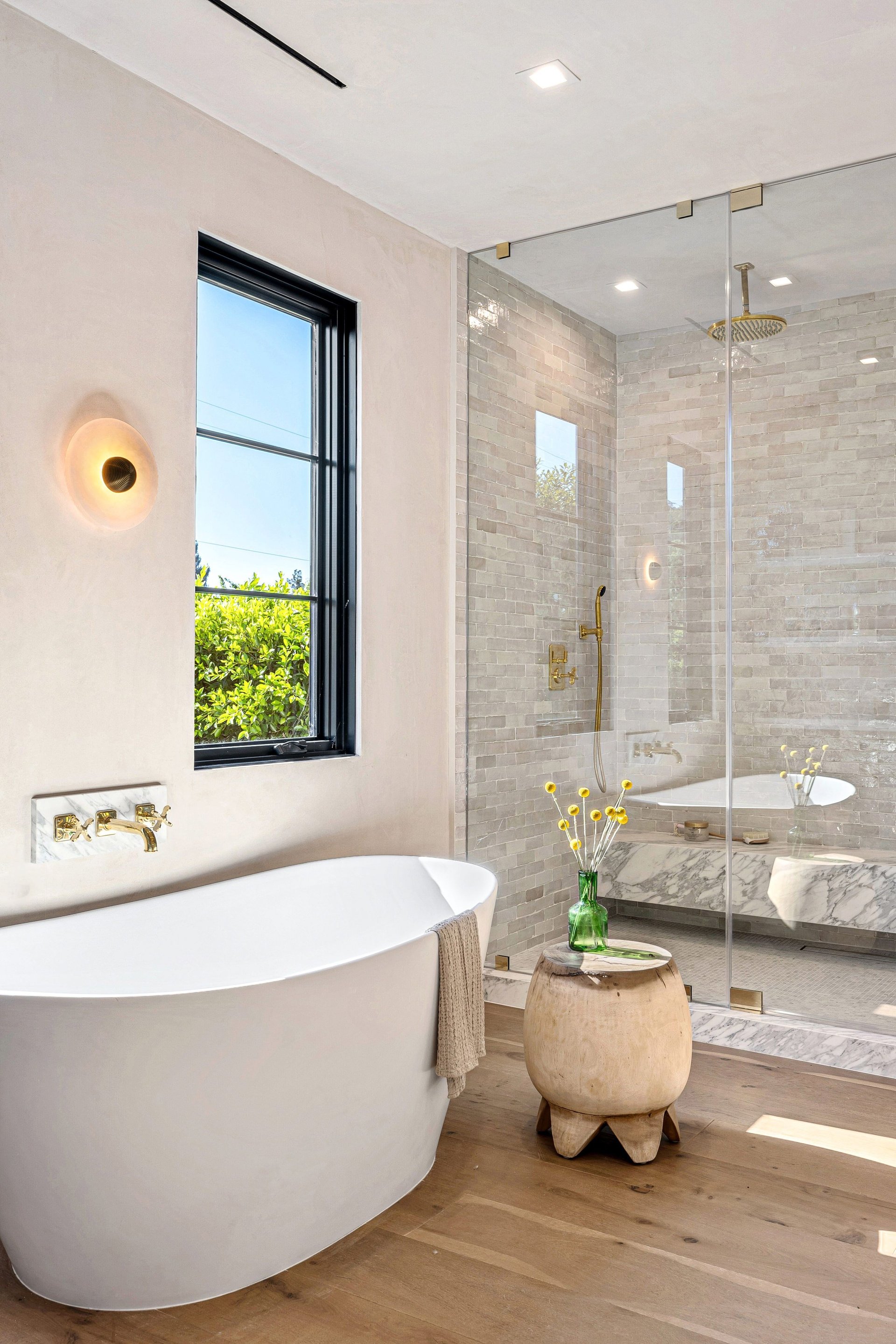 Modern bathroom with a freestanding tub, glass shower, and wooden accents, featuring natural light and elegant decor