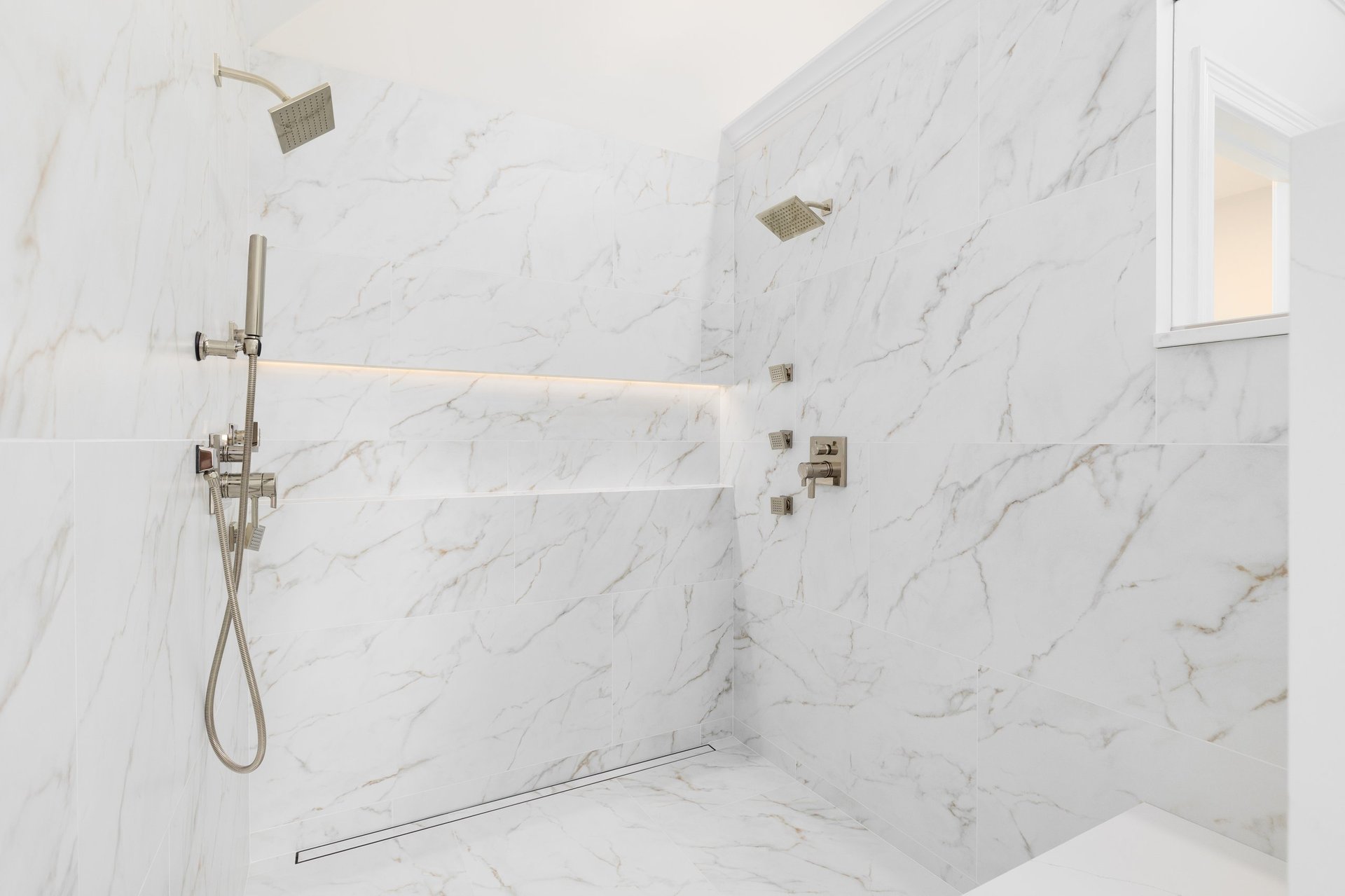 A large walk-in shower with large format white marble tiled walls, gold faucets, and a shelf with lighting