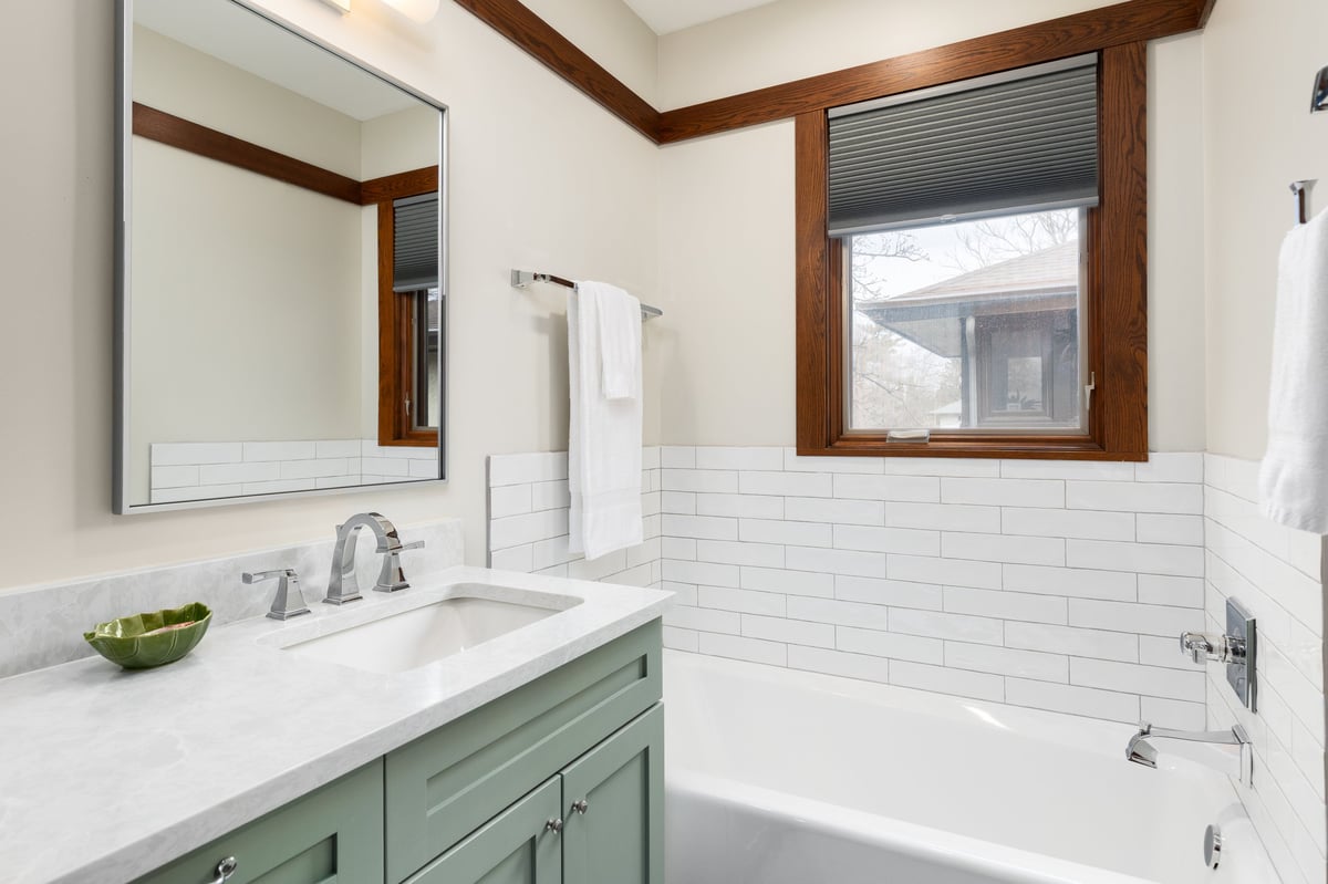 A bathroom sink detail with a chrome faucet on a white marble countertop, sage green cabinet, and a bathtub lined with white ceramic subway tiles.